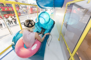 A young woman with blonde hair and a pink shirt sits in an inflatable pool toy at the top of a tube shaped waterslide in a store.