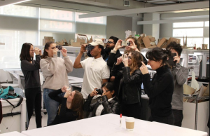 A group of 8 students hold up VR headsets standing around a table.