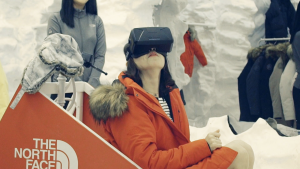 A woman sits in a reconstruction of a dog sled wearing a red coat and VR headset. She's using VR for retail in a North Face outdoor store.